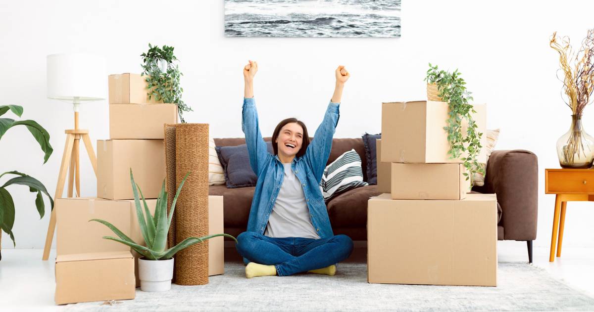 Vrolijke jonge vrouw zit blij op de grond tussen haar verhuisdozen in haar nieuwe huis